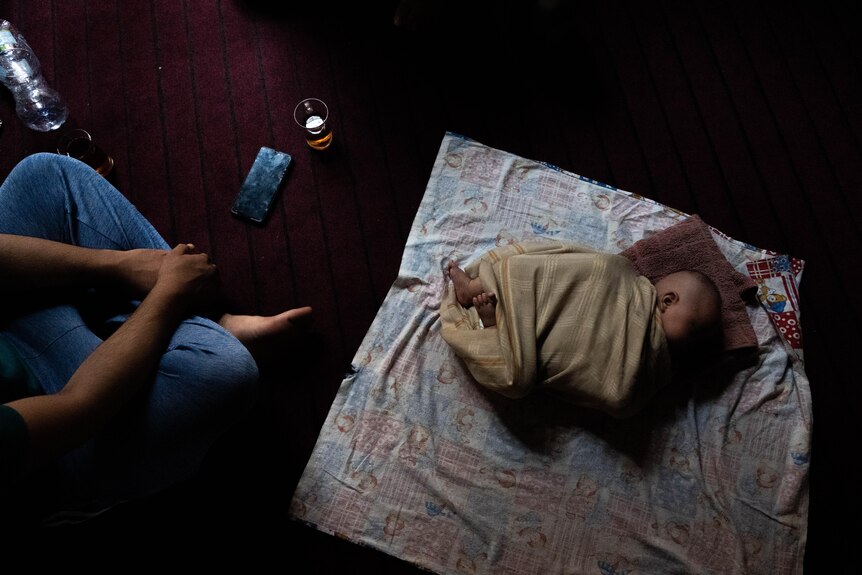 Taken from above, the photo shows a small baby wrapped in cloth sleeping on a plum-coloured carpeted floor. 