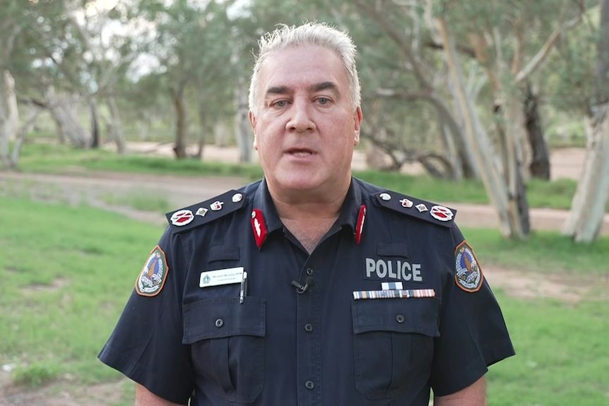 Northern Territory Police Commissioner stands in navy police uniform in front of open green field with gum trees.