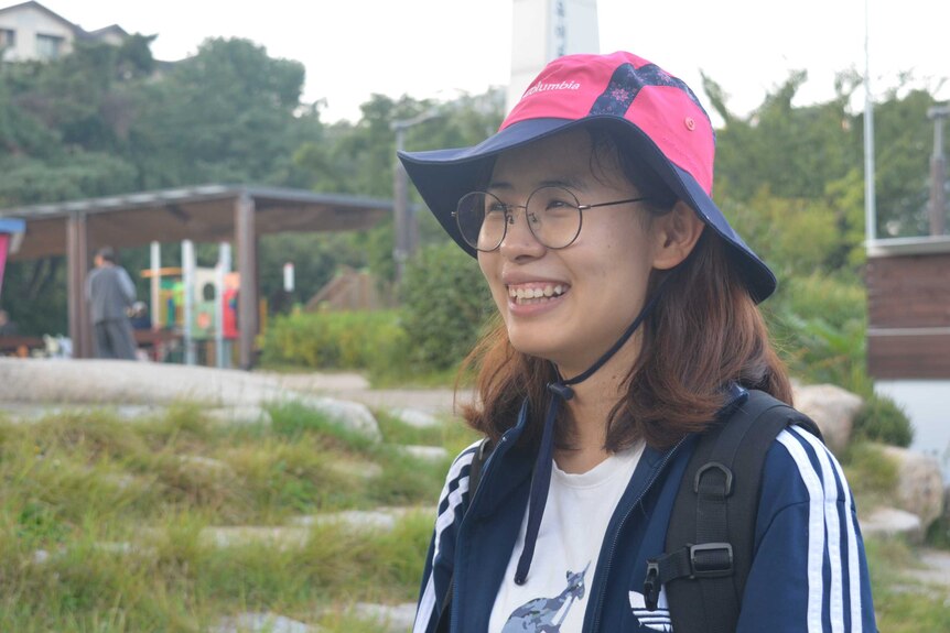 A woman in a pink bucket hat and glasses smiles and looks off into the distance.