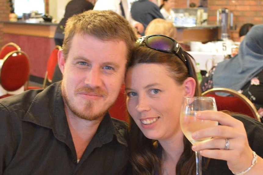 A man in a black shirt has his arm around a woman with sunglasses on her head, who is holding a glass of wine