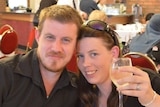 A man in a black shirt has his arm around a woman with sunglasses on her head, who is holding a glass of wine