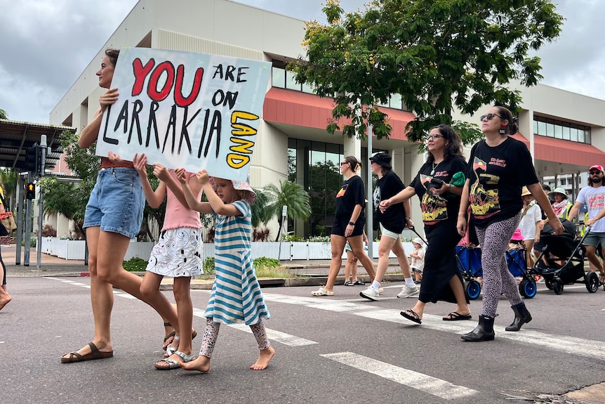 Three people hold a sign reading 'you are on Larrikia Land' as they walk with a big crowd.