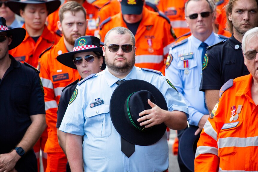 a man in a blue uniform holds his hat to his chest in respect as other people in various uniforms stand behind solemnly