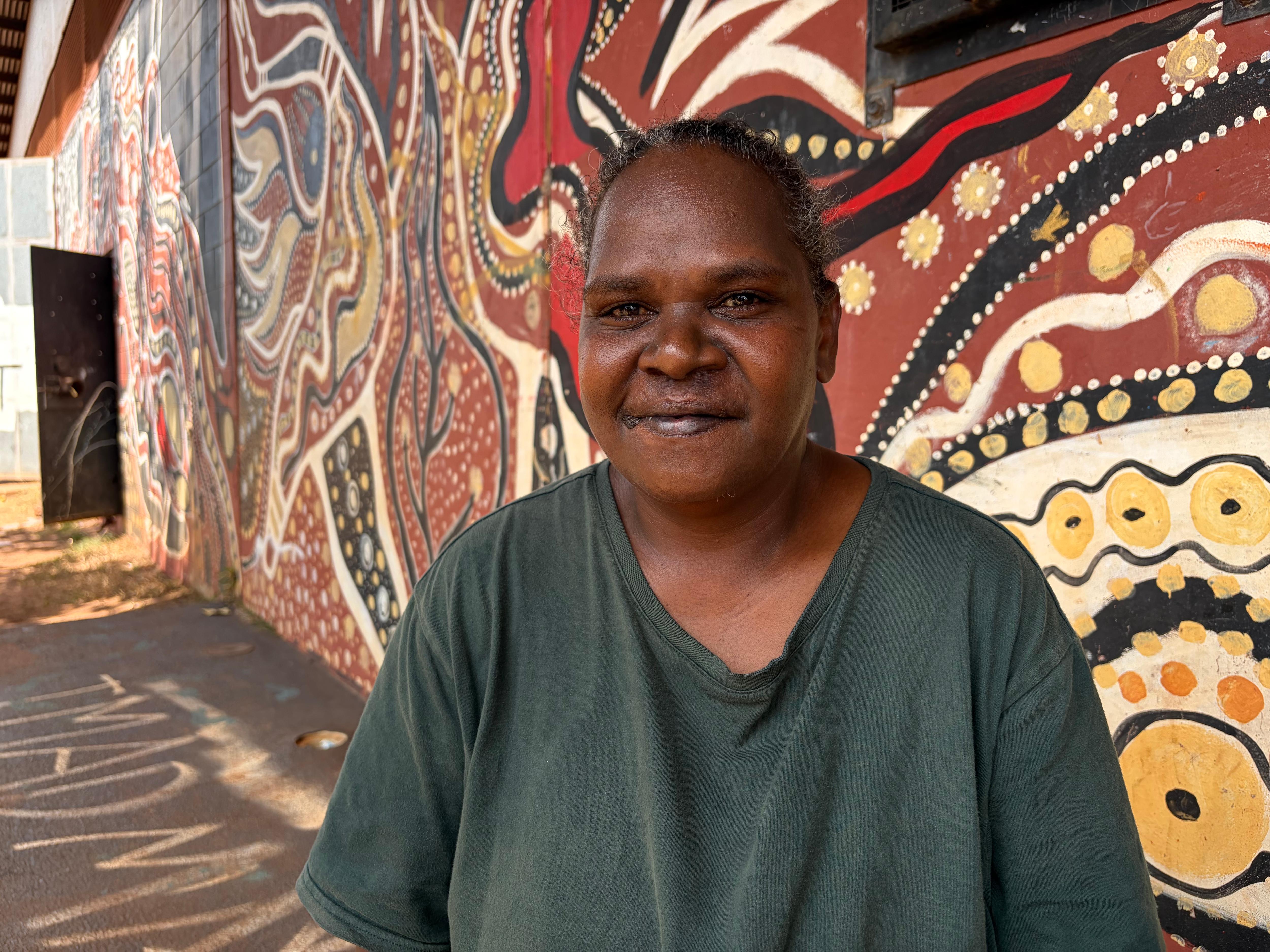 Indigenous woman Loretta Mills smiles while sitting in front of a wall covered in Aboriginal art dot painting.