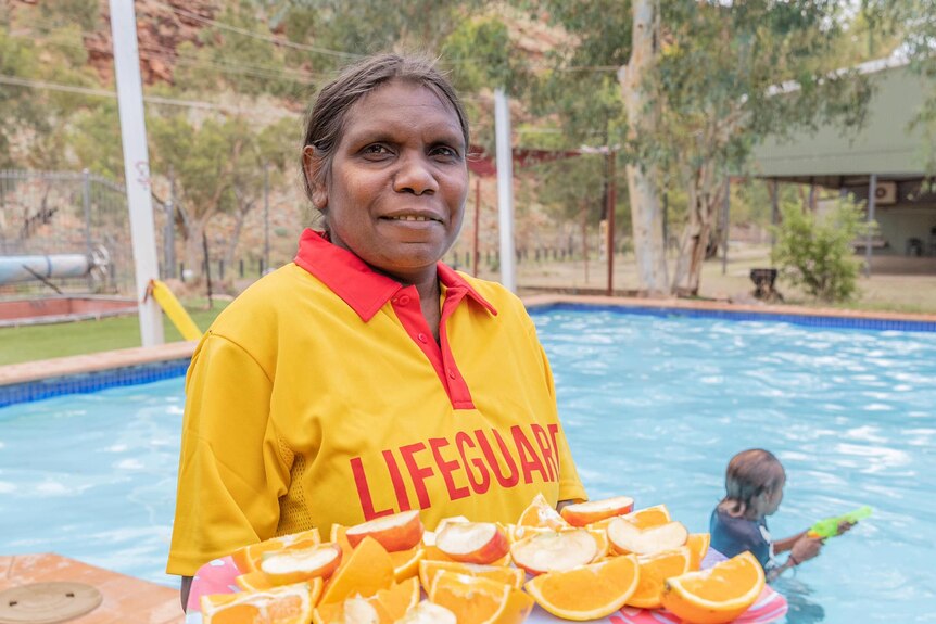 A woman wearing a lifeguard uniform holds a tray of cut-up oranges near a swimming pool.