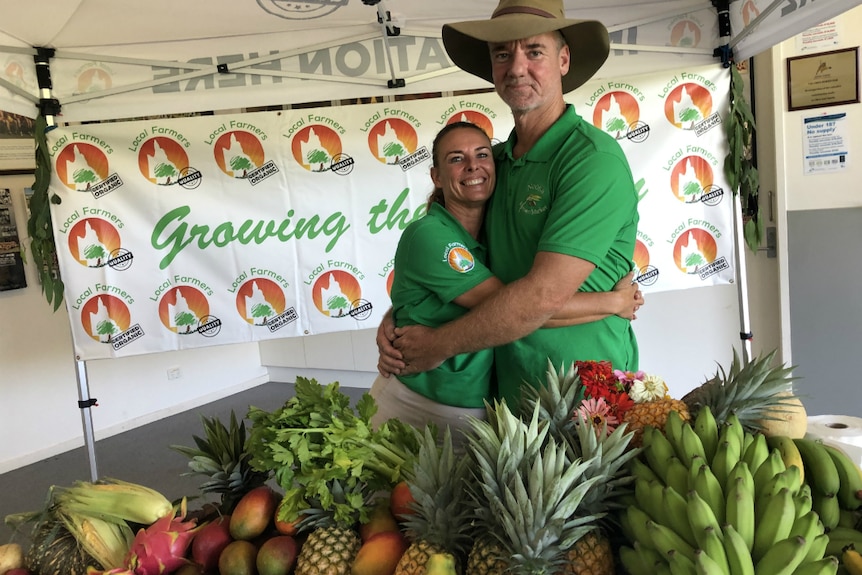 The couple hugs, standing behind fruit and vegetables on a table.