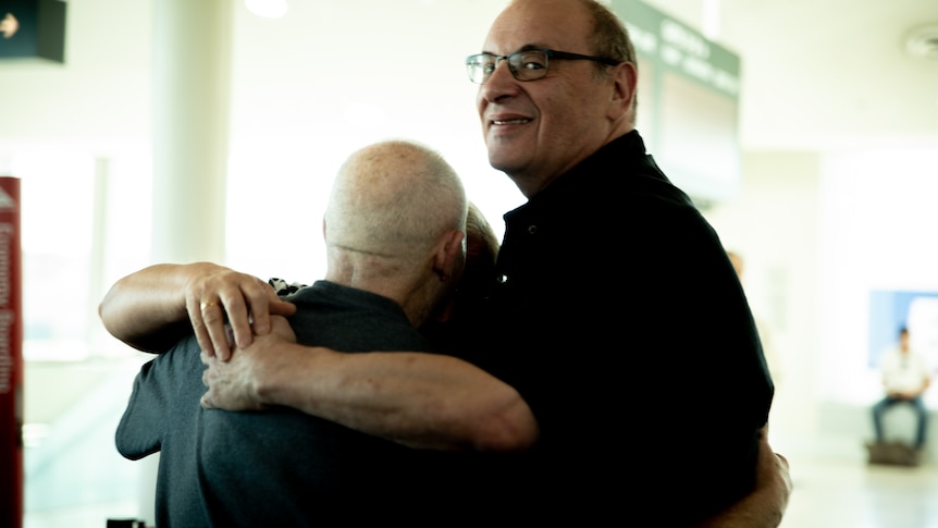 Two older men hug in an airport as one, who is wearing a black polo shirt and glasses, smiles towards the camera.