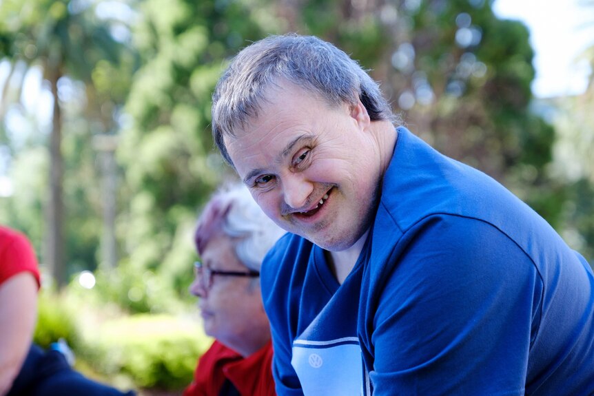Mark Deans wears a blue t-shirt and smiles at the camera while sitting in a group circle in a park on a sunny day
