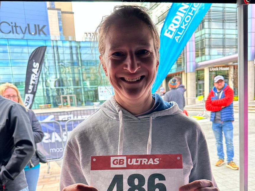 A woman in a white hoodie holds up a running bib number..