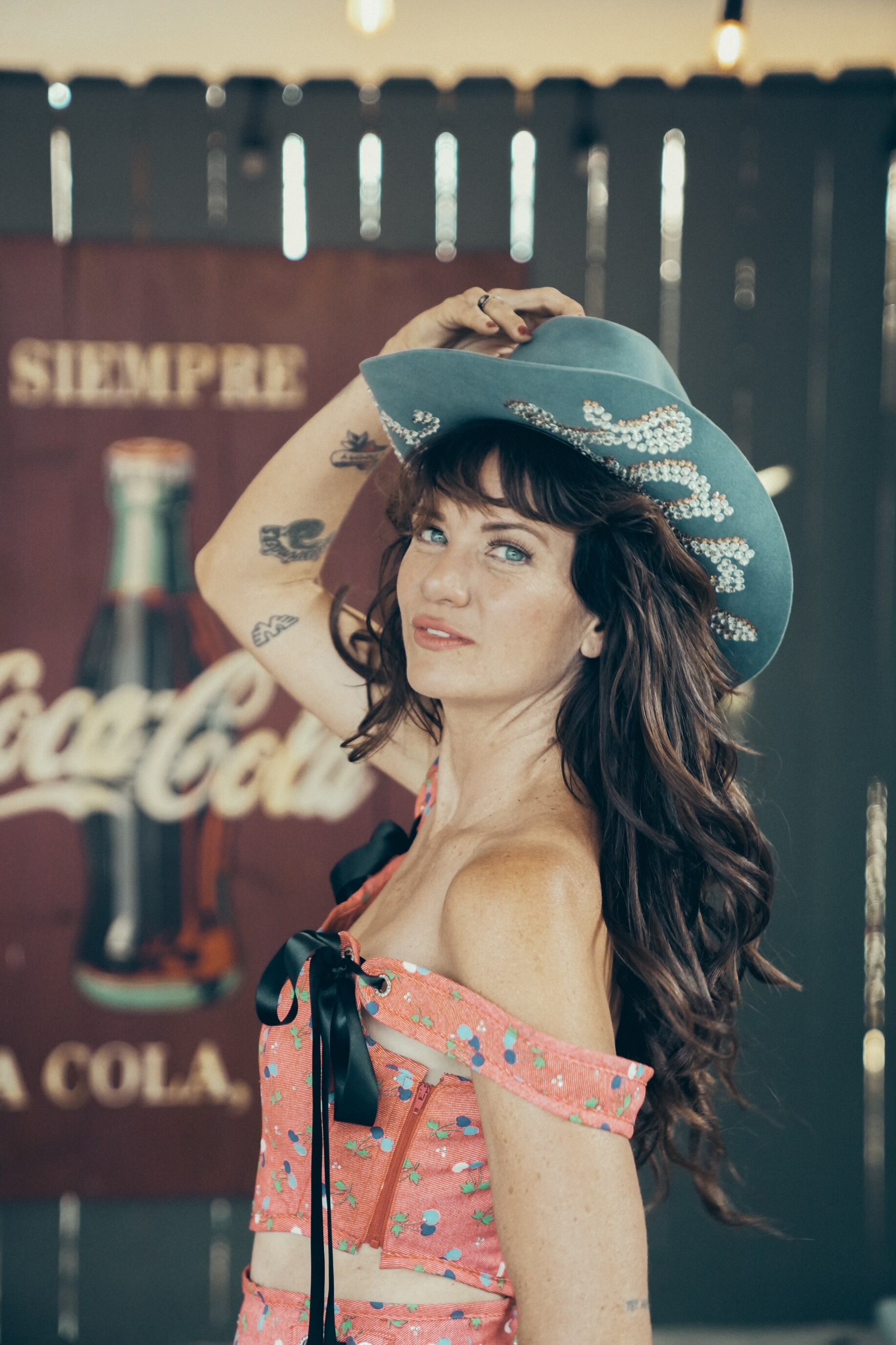 A woman holds onto the blue cowboy hat that's on her head. She stands beside a Coca Cola sign.