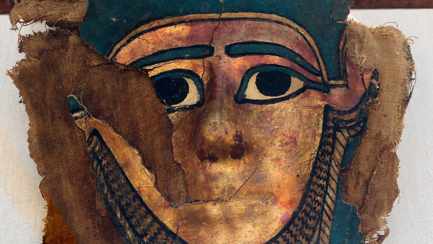 A broken gilded mummy mask is laid on a brown piece of cloth.