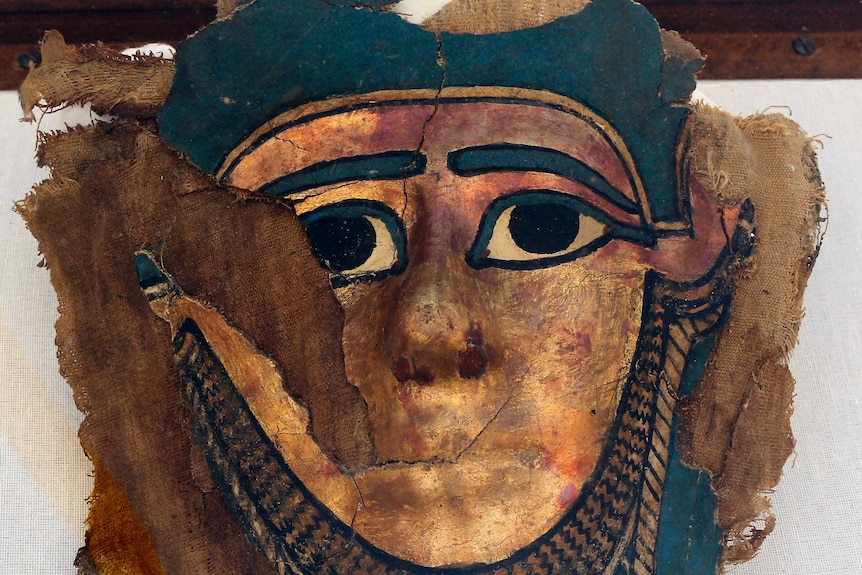 A broken gilded mummy mask is laid on a brown piece of cloth.