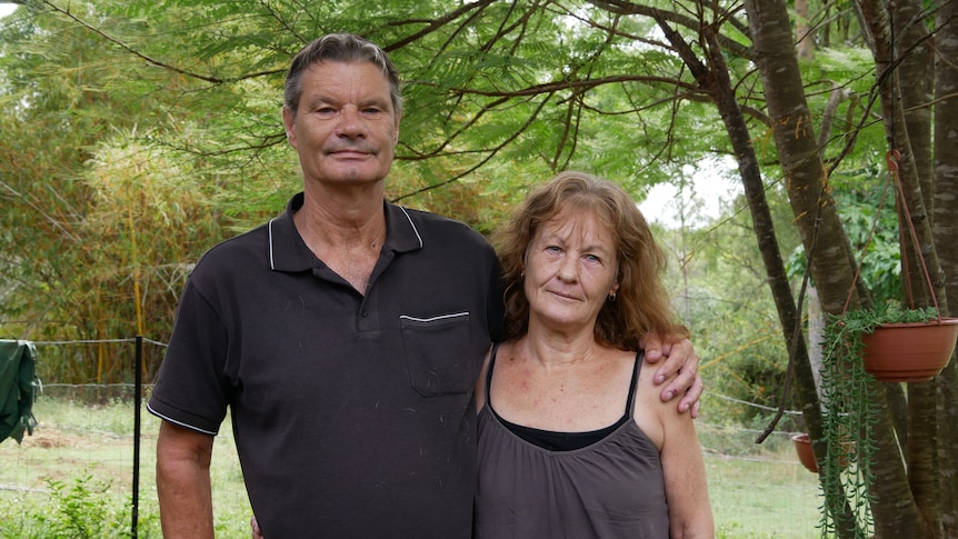A man and woman stand in front of a tree and look sternly at the camera
