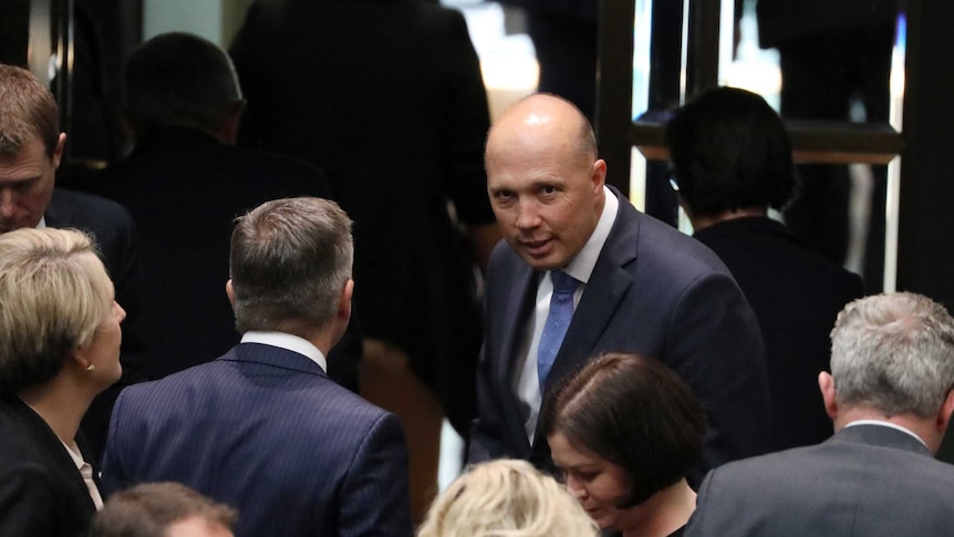 A man, dressed in a suit is surrounded by other professionally dressed people, glances up at the camera