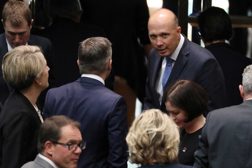 A man, dressed in a suit is surrounded by other professionally dressed people, glances up at the camera