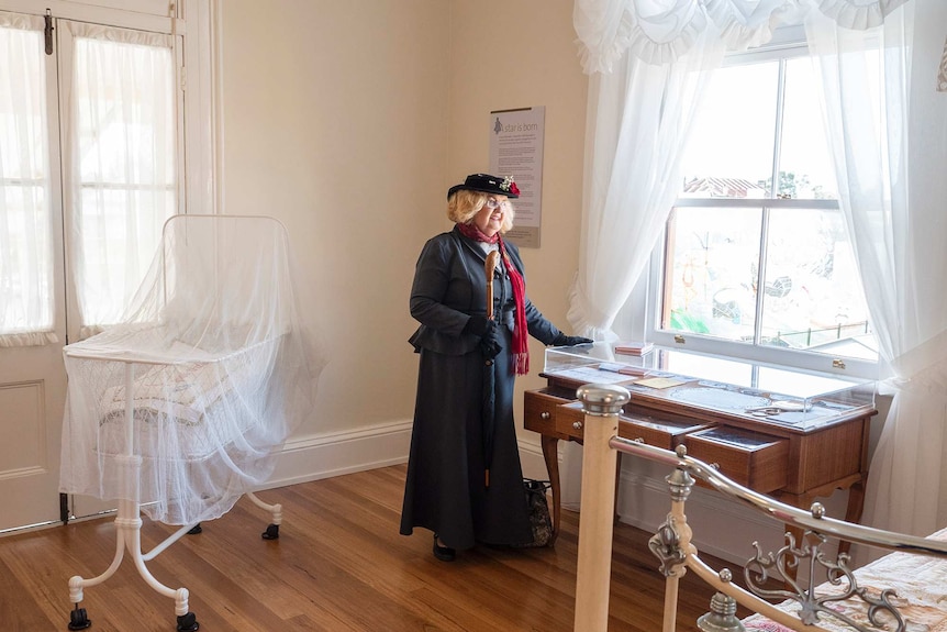 An older woman dressed in costume as Mary Poppins stands in a room featuring an antique crib on display.