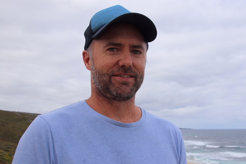 A man in blue shirt and cap stands over a beach with sky and water in the background.