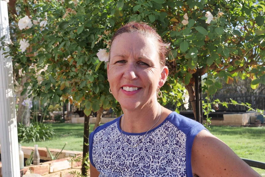 Woman with brunette hair in blue patterned top stands in front of white rose bush, smiling.