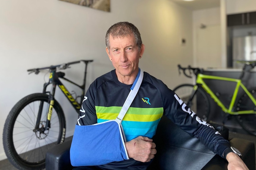 A man sits in a room with two bicycles. He wears a sling for a broken arm.