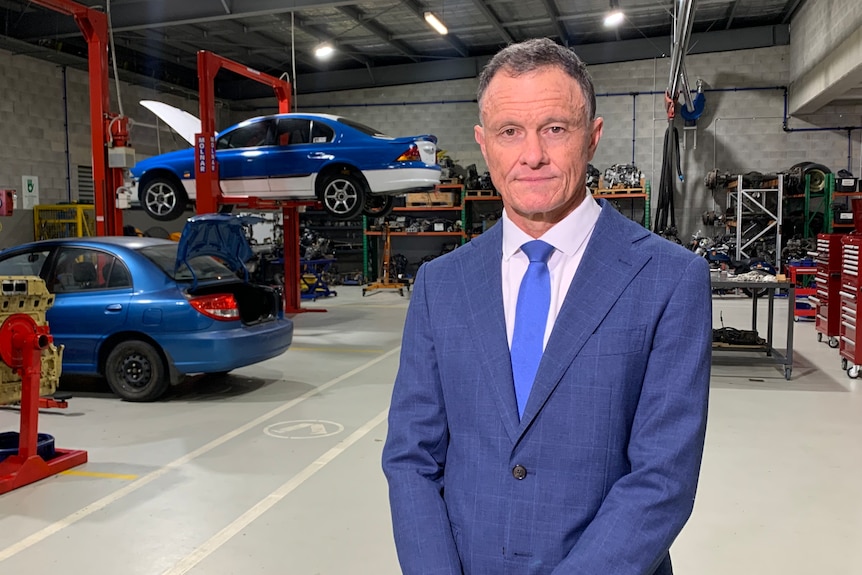 Man in suit standing in mechanic workshop with car on hoist in background