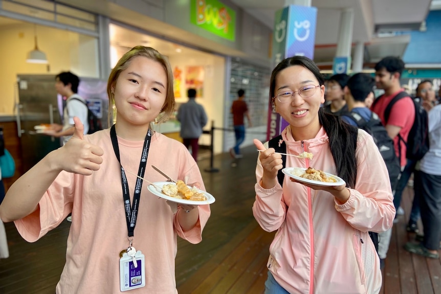 two young women holding a plate of food, one gives a thumbs up to the camera