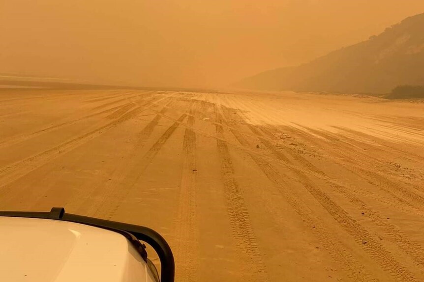 A car is seen to the left of the frame with a beach in front and a hill to the right. The sky is orange and hazy.