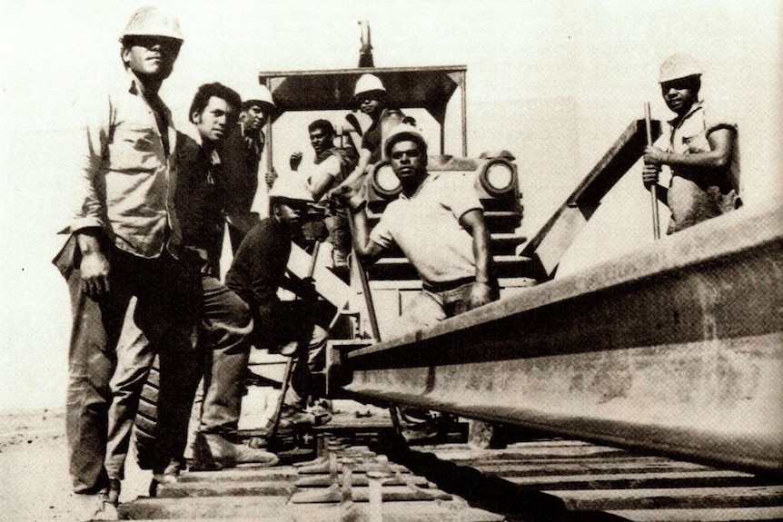 Fotografía en blanco y negro de un grupo de trabajadores varones de pie sobre una vía férrea.