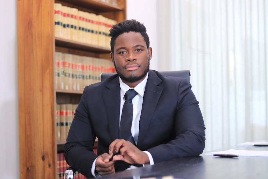 A man wearing a suit and tie sits in an office with leather-bound books in a book case behind him.