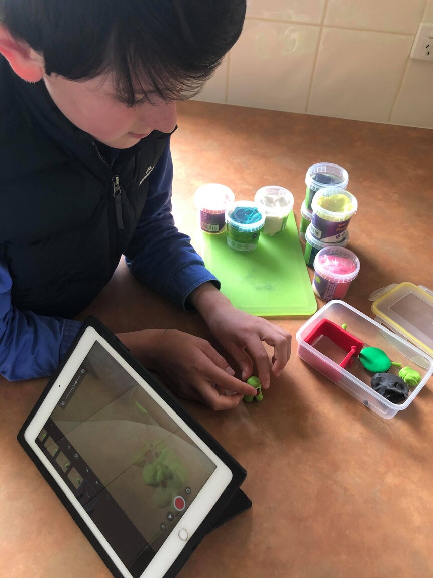 A boy leaning on at desk, with a tablet device and pots of coloured modelling compound around him as he builds a character