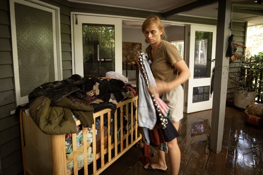 a woman sorts through clothes that are piled into a cot which is sitting on a verandah outside a house hit by flooding