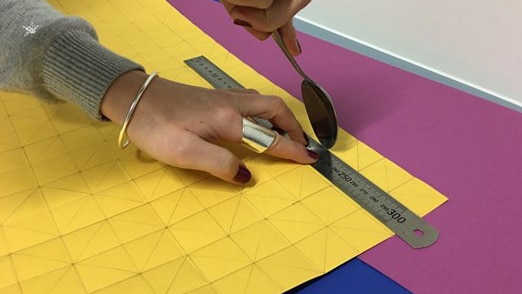A hand scores along a steel ruler as it lays on a paper with a grid pattern drawn on it, to form a crease in the paper