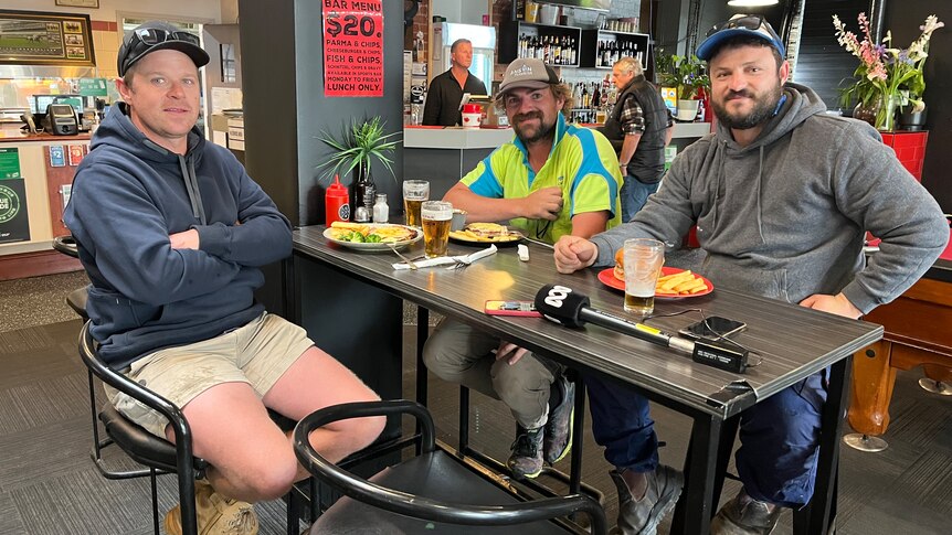 Three men wearing caps and hoodies sit around a table waiting for food in a bar.