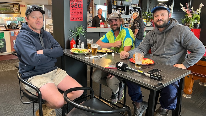 Three men wearing caps and hoodies sit around a table waiting for food in a bar.