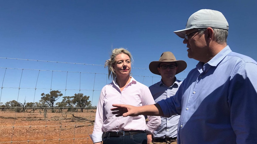 Scott Morrison in a cap gestures with one hand while talking to Bridget McKenzie and David Littleproud leaning against a fence.