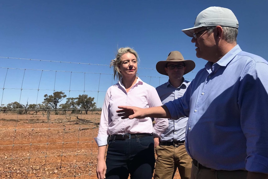 Scott Morrison in a cap gestures with one hand while talking to Bridget McKenzie and David Littleproud leaning against a fence.