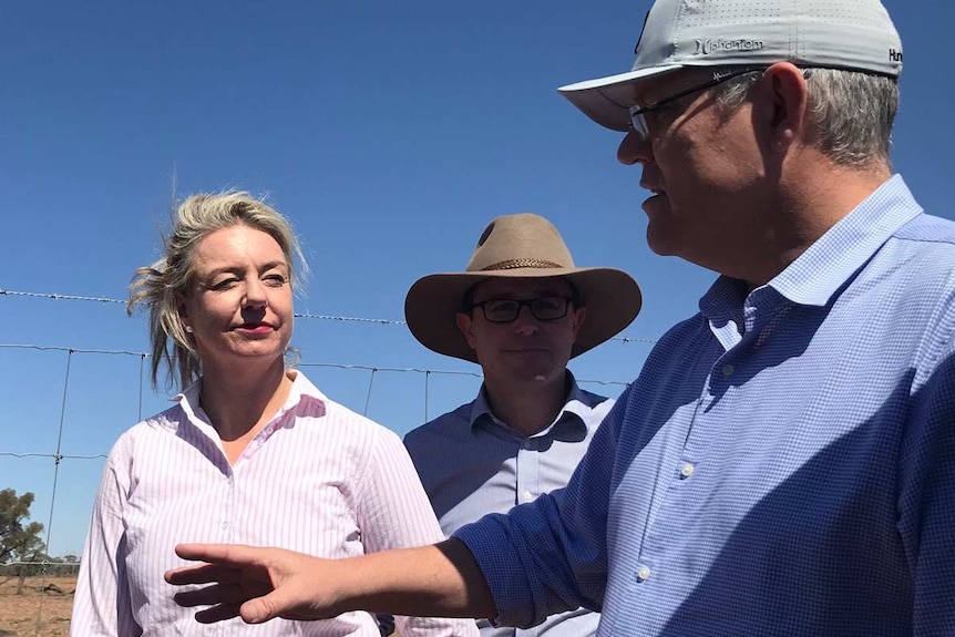 Scott Morrison in a cap gestures with one hand while talking to Bridget McKenzie and David Littleproud leaning against a fence.
