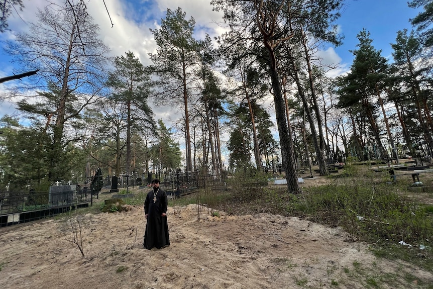 A priest stands on top of sandy ground in the middle of a forest cemetery. 