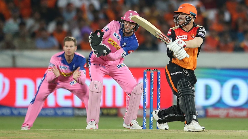 David Warner, wearing orange, plays a shot and looks over his right shoulder as Jos Buttler and Steve Smith stand behind in pink