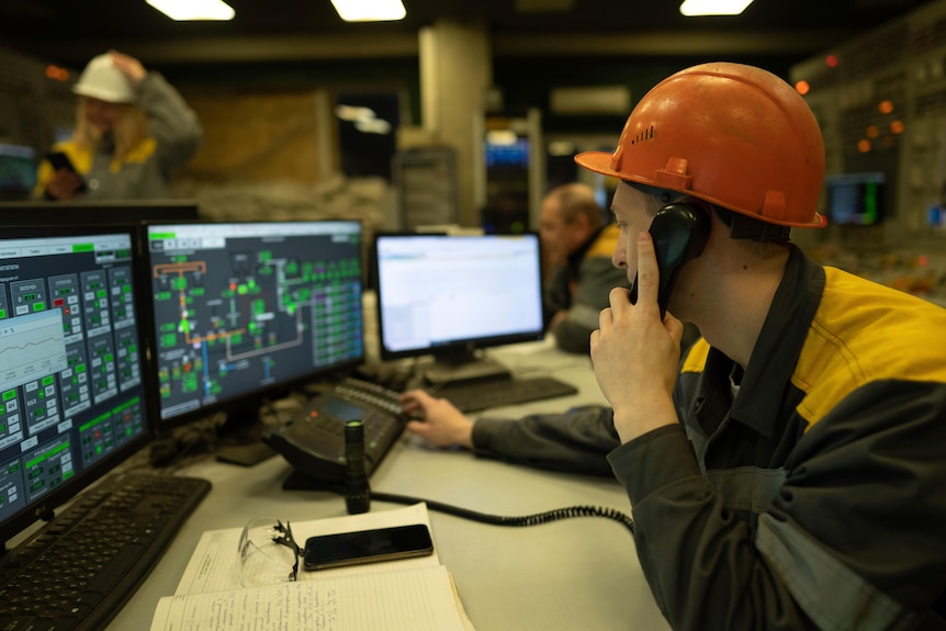 a power plant worker in a hard hat puts a phone receiver as he looks at computer monitor