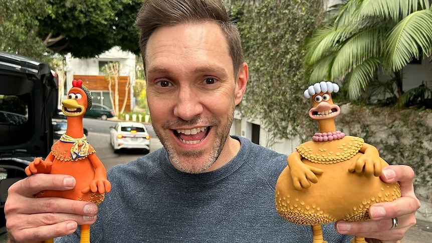 Andy Bailey, who has short hair and light stubble, grins holding two of the Chicken Run figurines.