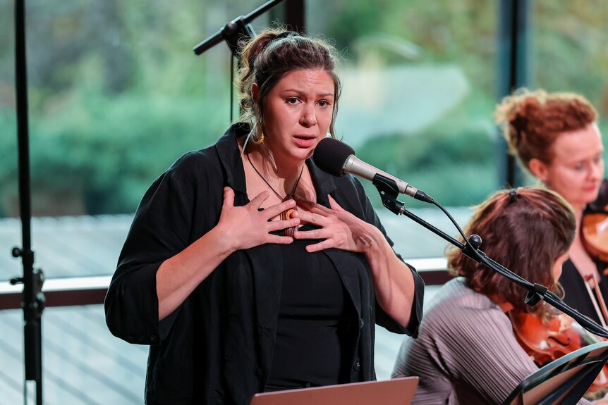 Young Yorta Yorta woman with brown hair pulled back, hands on chest, speaking into mic, on stage.