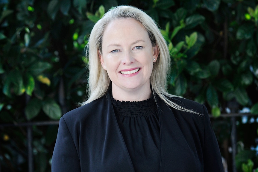 An image of Michell stewart, who has blonde shoulder-length hair, smiling, wearing black, in front of green tree leaves