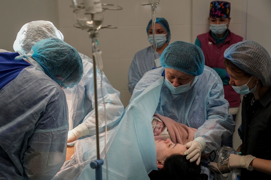 Medical workers hold a newborn child close to their mother's face, as others attend to the mother's body mid-surgery.