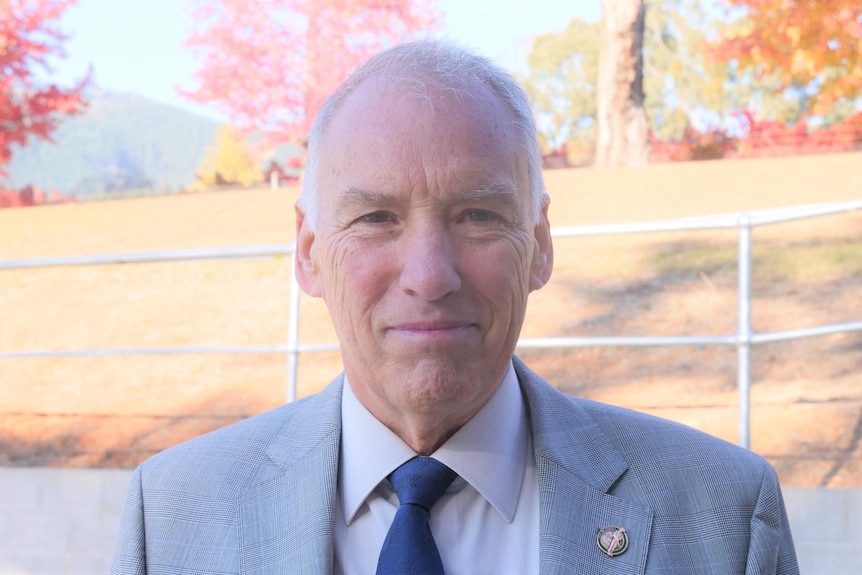 An older grey-haried, white man wearing a grey suit with autumn trees in the background