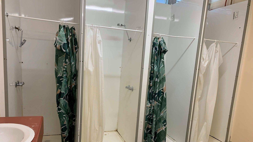 Four side-by-side shower cubicles with shower curtains. The floor in the bathroom is discoloured