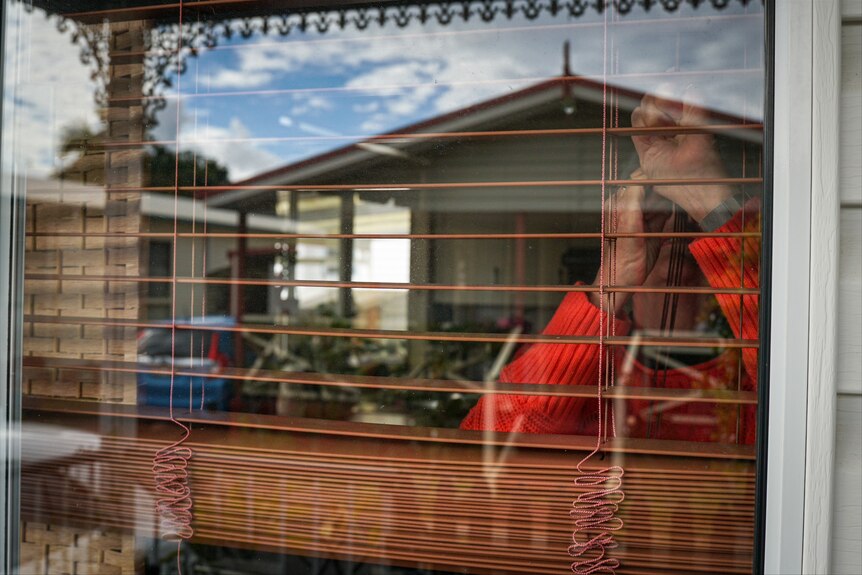 An older woman pulls up the blinds in her home, as seen from without.