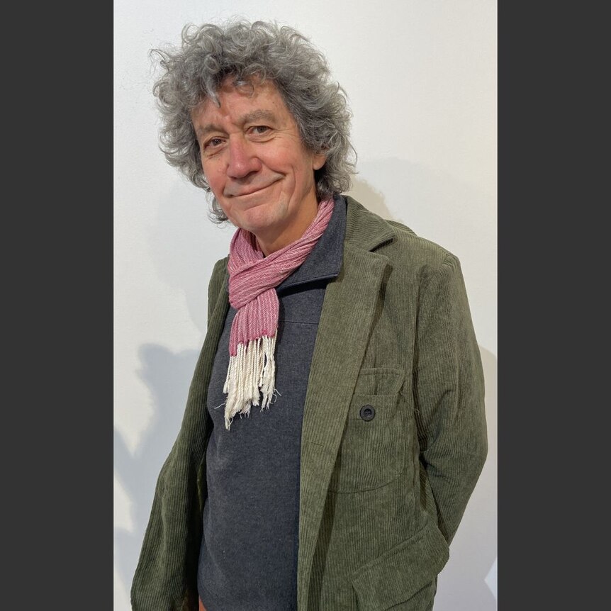 A man with grey, curly hair leans away from the camera, wearing a red and white scarf and a green blazer.