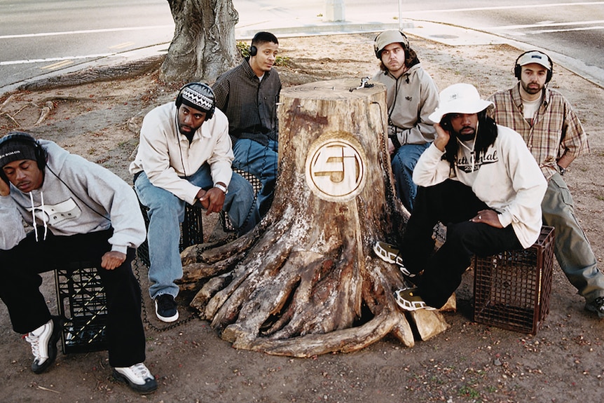 The six members of Jurassic 5 sit around a tree stump with headphones on, ignoring one another.