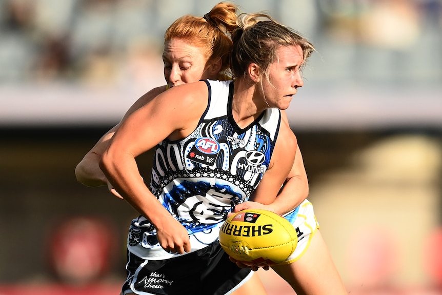 A Carlton AFLW player looks to her left as she tries to get a handball away with a Gold Coast opponent tackling her from behind.