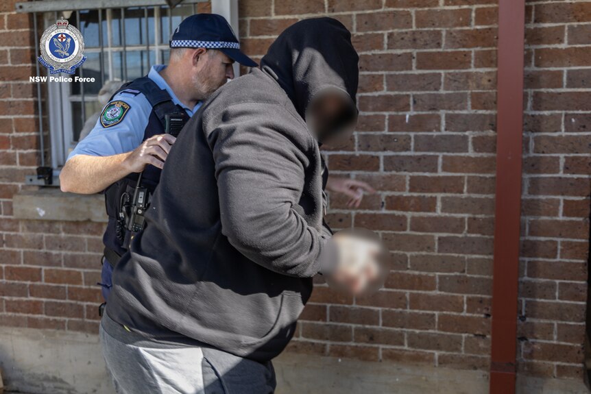 A man wearing a hooded jumper is walked towards a brick building by a police officer.